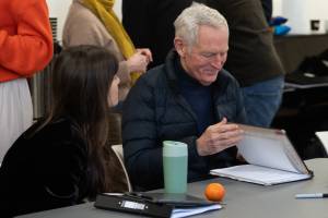 Two people reviewing scripts together at a table, with various items like a notebook, a water bottle, and an orange in front of them. The person on the right is smiling.