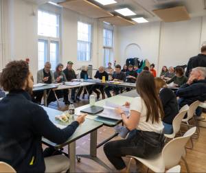 A group of actors sit around a large square of tables looking at scripts and engaging in friendly conversation.